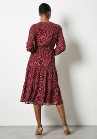 Vrouw die met haar rug naar de camera staat en een bordeaux lange mouwen midi-jurk met witte stippen en beige hoge hakken draagt.