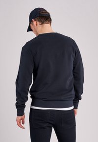 Mörk marinblå sweatshirt med långa ärmar, ribbade muddar och rundad halsringning. Bärs över en vit skjorta, med en matt yta och inga synliga mönster.