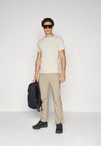 Šviesiai ruda trumpomis rankovėmis marškinėliai su mažu logotipu, smėlio spalvos kelnės, juodas kuprinė, juodi žygio batai ir tamsūs saulės akiniai.