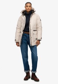 Kobieta w beżowej zimowej kurtce z kapturem, granatowym swetrze z golfem, niebieskich dżinsach z podwiniętymi nogawkami, brązowym plecionym pasku i brązowych butach na sznurówki.