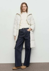 Mujer vistiendo un abrigo acolchado largo blanco, suéter crema, jeans de pierna ancha azul oscuro y zapatos mocasines color beige, de pie contra una pared clara.