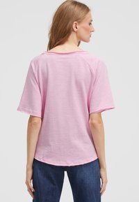 Woman with light brown hair wearing a loose pink short-sleeve shirt and dark blue jeans, facing away against a plain light background.