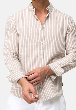 Mand justerer manchet på beige og hvidt vertikalt stribet skjorte med knapper, iført hvide bukser, mod en ensfarvet baggrund.