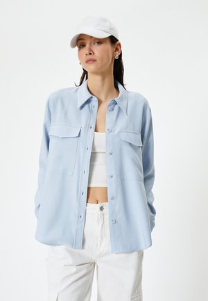 Camisa azul claro de botones con dos bolsillos en el pecho, fabricada en tejido ligero y de corte relajado, llevada sobre una camiseta blanca corta y pantalones blancos.