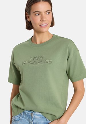 Femme portant un t-shirt vert clair à manches courtes avec le texte "LINES IN THE SAND UNDER DESERT SKIES", associé à un jean bleu clair.