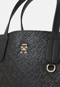Zwarte handtas gemaakt van gestructureerd synthetisch materiaal, met een geometrisch patroon, twee handvatten en een gouden logoaccent.