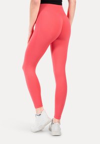 Persoon die koraalroze textuur legging en witte sportschoenen draagt, gezien van achteren tegen een effen lichte achtergrond.