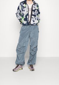 Puszysta granatowa kurtka z abstrakcyjnymi wzorami, zapinana na zamek, szare spodnie cargo o lśniącej fakturze oraz lekkie, fioletowe buty do biegania.