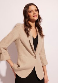 Beżowa lniana marynarka o dopasowanym kroju, z klapami, pojedynczym zapięciem na guzik i podwiniętymi rękawami, zestawiona z czarną sukienką z prążkowanej tkaniny.