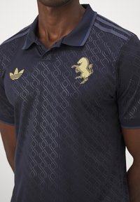 Marineblaues Poloshirt mit strukturiertem Rautenmuster, goldenem Löwenlogo und Adidas-Streifen auf den Schultern. Der Stoff wirkt glatt und leicht.