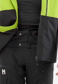 Veste étanche noire avec une partie supérieure vert vif, dotée d'une fermeture éclair, de boutons-pression et d'un tissu texturé. Logo sur le pantalon.