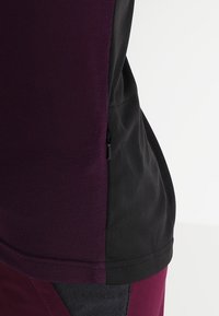 Kurzärmliges sportliches Shirt in Dunkelpurpur und Schwarz. Aus atmungsaktivem Mesh-Material mit seitlichem Reißverschluss. Insgesamt glatte Textur.