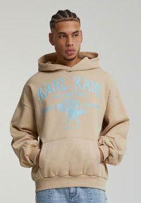 Felpa beige con cappuccio e grafica blu con testo "KARL KANI" e design di un'aquila. Presenta una tasca frontale e polsini e orlo a coste.