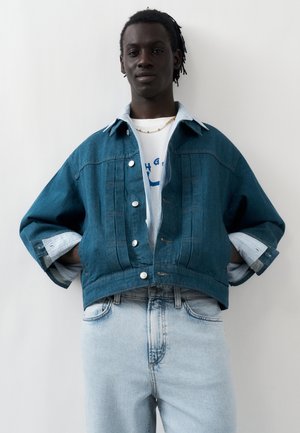 Homme avec de courtes dreadlocks portant une veste en denim bleue superposée sur un t-shirt blanc à motif graphique et un jean bleu clair, debout les mains sur les hanches.