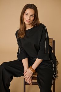 Femme aux longs cheveux bruns portant une tenue noire avec bordure blanche, assise sur une chaise en bois sur fond beige, les mains jointes avec un bracelet marron.