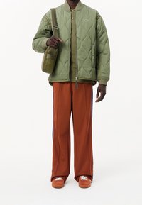 Vadderad grön jacka med dragkedja, olivfärgad skjorta, rostfärgade vida byxor med blå ränder och bruna sneakers med vita snörningar. Brun axelremsväska.