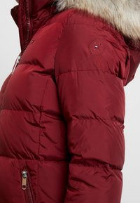 Giacca puffer bordeaux con cappuccio in pelliccia sintetica, design trapuntato, tasca laterale con zip e un logo rotondo sulla manica superiore.