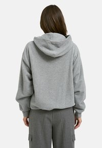 Person mit langen braunen Haaren, die einen lockeren grauen Hoodie und graue Hosen trägt, von hinten vor einem schlichten weißen Hintergrund gezeigt.