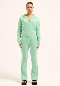 Tuta in velour verde menta con felpa con zip e pantaloni con cordino. Presenta dettagli in oro e una texture morbida, abbinata a sandali con plateau.