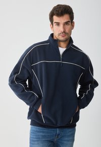 Sudadera de forro polar azul marino con cremallera de cuarto de vuelta y acentos en blanco. Presenta mangas largas, un cuello alto y bolsillos frontales. Tejido texturizado.