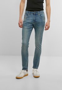 Šviesiai mėlyni džinsai su siauru siluetu, turintys penkias kišenes, šiek tiek išblukusį paviršių ir subtilius siuvinėjimo akcentus. Derinami su baltais sportbačiais.