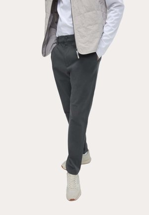 Mann trägt dunkelgraue Hose, weiße Turnschuhe, weißes Langarmhemd und beige gesteppte Weste, steht mit Händen in den Taschen.