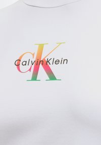 Logo Calvin Klein avec de grandes lettres "cK" superposées en dégradé orange, rose et vert sur un fond en tissu blanc.