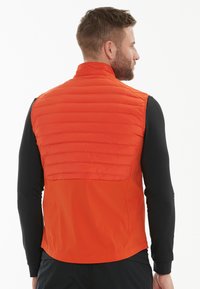 Orange vadderad väst med quiltad textur, hög krage och en kontrasterande svart långärmad skjorta under. Slät yta, avslappnad design.