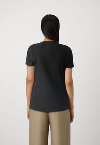 Žena s dlouhými tmavými vlasy, oblečená v jednoduchém černém tričku s krátkými rukávy a béžových kalhotách, stojící zády k bílé zdi.