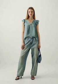 Young woman wearing teal sleeveless blouse with ruffles, wide-leg faded jeans, black heeled sandals, and holding a blue shoulder bag, standing.