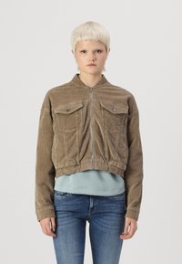 Kuttet kordjakke i khaki med glidelås foran, to brystlommer og ribbestrikkede mansjetter, båret over en lys blå genser og jeans.