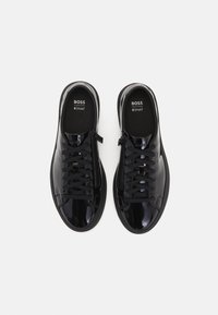 Μαύρα παπούτσια αθλητικά από γυαλιστερή δερματίνη με σχέδιο με κορδόνια, υφασμάτινες μαύρες λεπτομέρειες και λογότυπο στην εσωτερική σόλα. Διαθέτουν πλαϊνό φερμουάρ για εύκολη εφαρμογή.