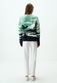 Pull en tricot avec un motif de paysage de montagne en vert, blanc et marine, présentant des rayures horizontales sur les manches et un col rond.