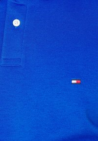 Blå polo-tröja med texturerad tyg, tvåknapps-knäppning och en liten broderad logotyp i röd, vit och blå på vänstra bröstet.