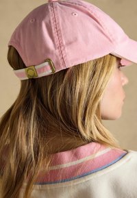 Persona rubia con una gorra de béisbol rosa con correa ajustable y un cuello de color claro con rayas rosas y azules.