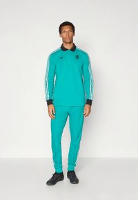 Σετ φόρμας τιρκουάζ με μακριά μανίκια και παντελόνι στο ίδιο χρώμα. Διαθέτει μαύρο γιακά, τρεις λευκές ρίγες στα μανίκια και το παντελόνι, καθώς και λογότυπα.