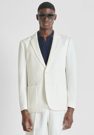 Hombre con blazer blanco extragrande y pantalones a juego, camisa azul marino y grandes gafas de sol oscuras, posando contra un fondo neutro.