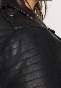 Black leather jacket with quilted texture on the shoulder, featuring snap button closure and smooth finish. Side profile view.