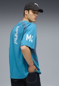 Blå t-shirt med kort ärm och horisontella ränder, som har "PUMA"-logotypen och texten "M4" i vitt. Bärs med mörka jeans och en svart keps.