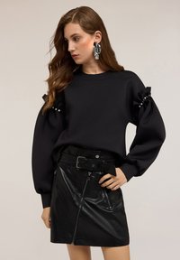 Donna con lunghi capelli castani che indossa un top nero a maniche puff con dettagli in perle e una gonna nera di pelle a vita alta con cintura.