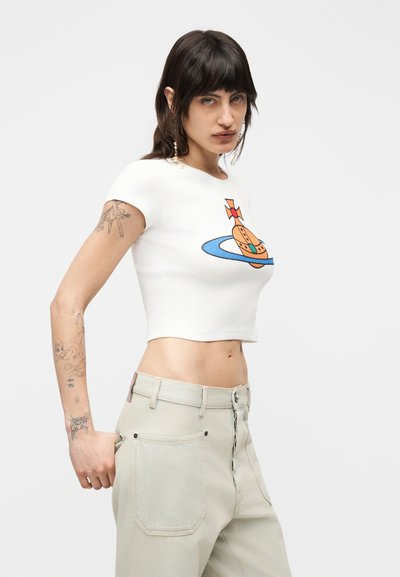 Γυναίκα με σκούρα μαλλιά που φοράει λευκό κοντό μπλουζάκι με πορτοκαλί και μπλε γραφικό και ανοιχτόχρωμο παντελόνι με ψηλή μέση, σε απλό φόντο.