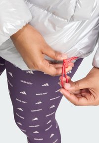 Sivi pufasti kaput s elastičnom crvenom šnrom koja se podešava. Ljubičaste tajice s ponavljajućim uzorkom bijelog loga "MOON BOOT".