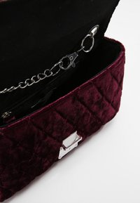 Bolso de mano de terciopelo acolchado en color burdeos con un cierre plateado y correa de cadena; presenta un interior negro con un bolsillo con cremallera y un sujeta llaves.