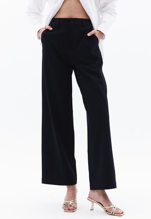 Zwarte wijde broek met een gladde textuur, voorzakken en een knoopsluiting. Gecombineerd met gouden sandaaltjes met hakken.