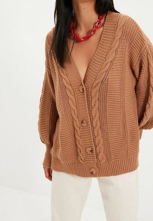 Frau trägt eine beige grob gestrickte Strickjacke mit Knöpfen, Zopfmuster, weiße Hose und eine grobe rote Ketten-Halskette.