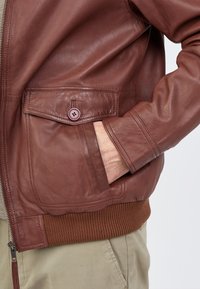 Braune Leder-Bomberjacke mit strukturiertem Finish, Frontreißverschluss und einer knöpfbaren Brusttasche. Mit gerippten Strickbündchen und Saum.