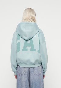 Felpa azzurra chiara con un grande logo verde "GAP" sulla schiena, con cappuccio regolabile e polsini a coste. Indossata con jeans larghi.