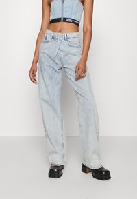 Persona que lleva puestos unos pantalones de mezclilla de tiro alto y pierna ancha de color azul claro, un top de mezclilla recortado con ribete negro y zapatos de plataforma negros, de pie contra un fondo blanco.