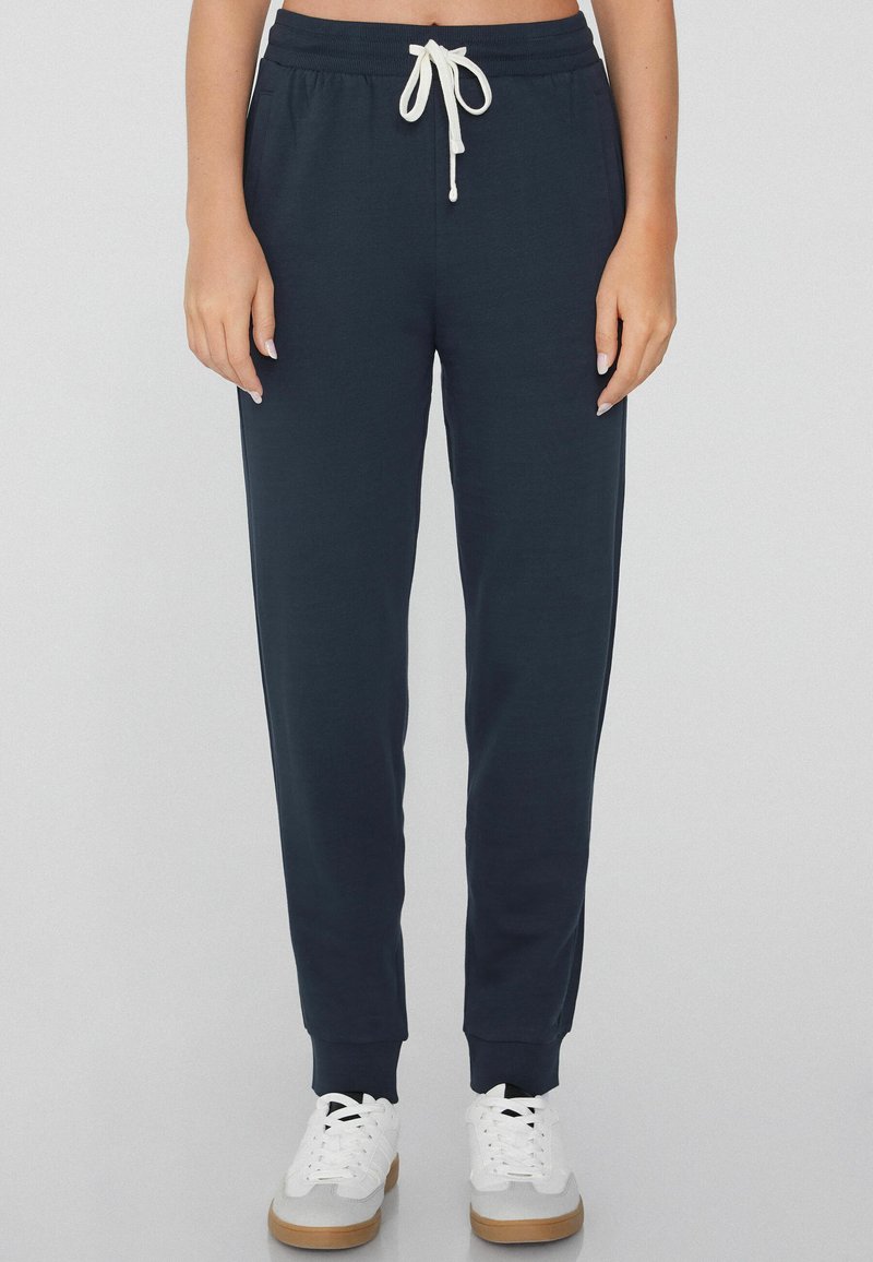 Pantalones jogger azul marino hechos de tela suave, con una cinturilla elástica y cordón blanco, además de puños acanalados. Combinados con zapatillas blancas.