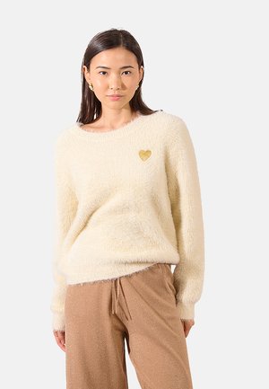 Maglione fluffy color crema con collo rotondo e maniche lunghe; presenta un piccolo cuore d'oro come decorazione sul petto.